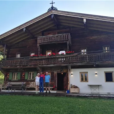 A traditional two-story wooden house in a picturesque landscape. Three people are standing on the porch in front of the house.