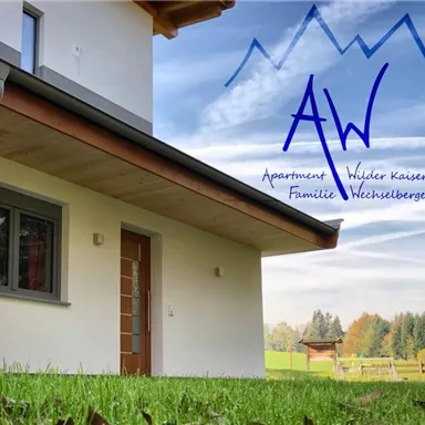 A modern apartment building surrounded by green grass and trees. In the background, gentle hills and a clear sky can be seen.