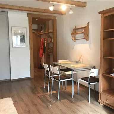 A bright room with a table and white chairs. In the background, there is a shelf and a wardrobe.