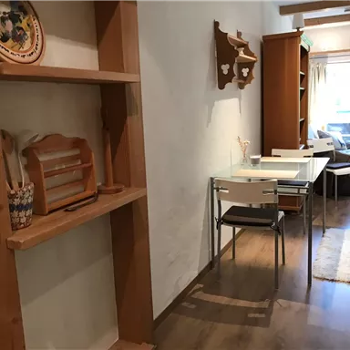 A bright hallway with wooden shelves and a glass table. In the background, chairs and a cozy seating area are visible.