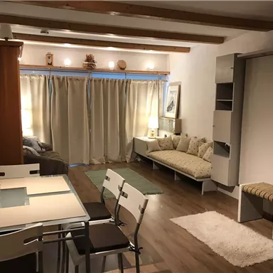 A cozy living room with a dining table and chairs. The room has light curtains, a sofa area, and warm lighting.