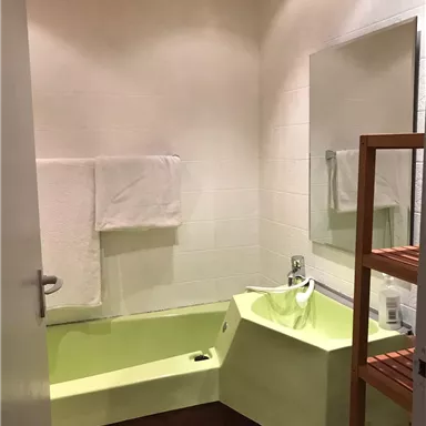 A modern bathroom with a green bathtub and a large mirror. The room contains white towels and a wooden shelf.