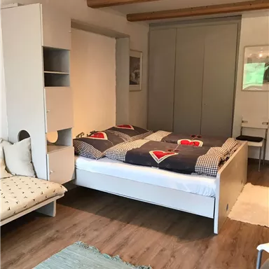 A cozy bedroom with a double bed and colorful bedspreads. The room includes a shelf, a couch, and a table with chairs.