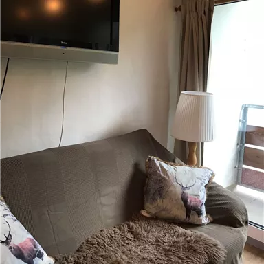 A cozy living room with a brown sofa and decorative pillows. A TV is mounted on the wall and there is a lamp next to a window.