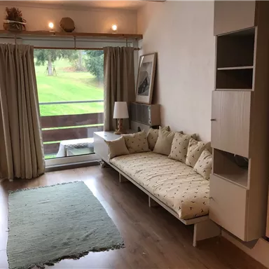 A bright living room with a cozy bench and a large window. Outside, a green meadow is visible.