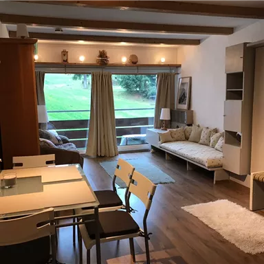 A cozy living room with a dining table and chairs. Large windows provide a view of the green landscape.