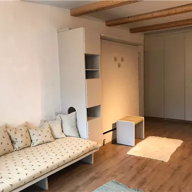 A cozy living space with a comfortable sofa and bright walls. It includes a small table and a wardrobe as well as a soft rug.