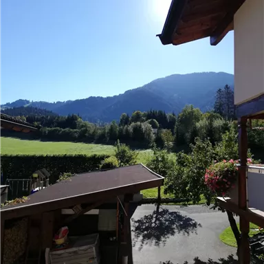 A picturesque landscape with mountains in the background and a clear sky. In the foreground, there is a wooden building and plants.