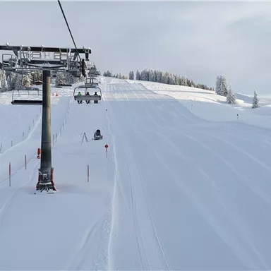 Eine Skipiste mit frischem Schnee und einer Seilbahn im Vordergrund. Die Bäume sind schneebedeckt und der Himmel ist leicht bewölkt.