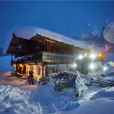 A snow-covered cabin at night, surrounded by a lot of snow. A snow plow is visible, clearing the road.