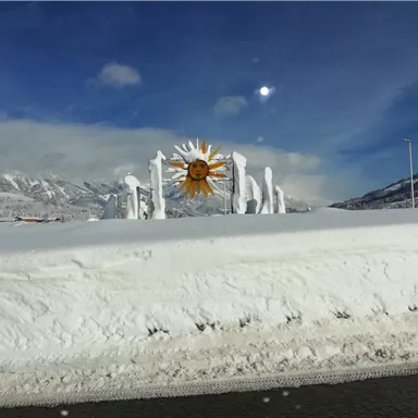 A snowy landscape with a colorful sun symbol in the center. The mountains can be seen in the background, and the sky is partially cloudy.