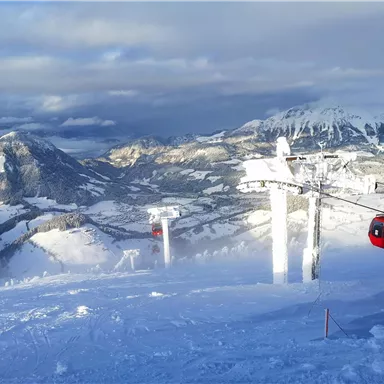 Eine verschneite Berglandschaft mit Skiliften und Gipfeln im Hintergrund. Der Himmel ist teilweise bewölkt und die Aussicht ist atemberaubend.