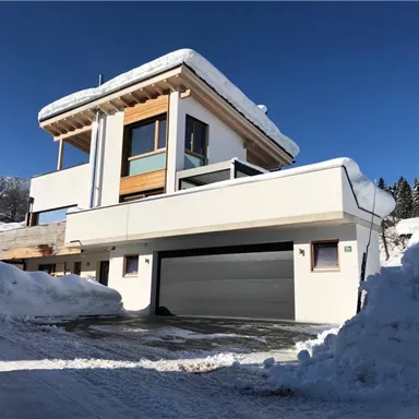 A modern house in the snow with a clear blue sky. The surroundings are wintry and the facade is partially covered with snow.