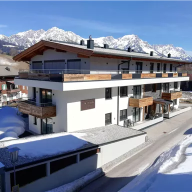 A modern building in the snow with balconies and large windows. In the background, impressive mountains and blue sky can be seen.