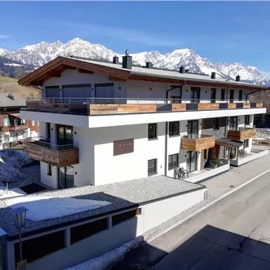 A modern building with balconies and wooden decorations. In the background, snow-covered mountains can be seen.