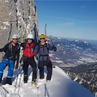 Drei Skifahrer stehen auf einem schneebedeckten Gipfel mit Blick auf die Berge. Der Himmel ist klar und blau.