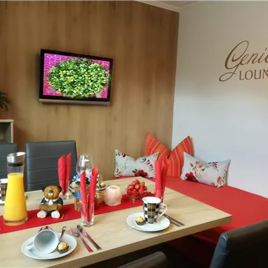Ein gemütlicher Essbereich mit einem Tisch, dekoriert mit Geschirr und Getränken. An der Wand steht der Schriftzug "Genießer Lounge" und ein Fernseher zeigt eine grüne Pflanzenansicht.