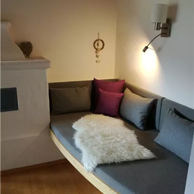Ein gemütlicher Sitzbereich mit grauen Kissen und einem weißen Fell. Die Wand ist hell, und eine Lampe sorgt für sanftes Licht.