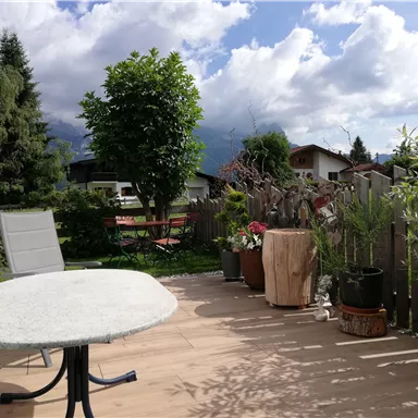A terrace with a table and a chair, surrounded by plants and trees. The sky is partly cloudy with some sunshine.