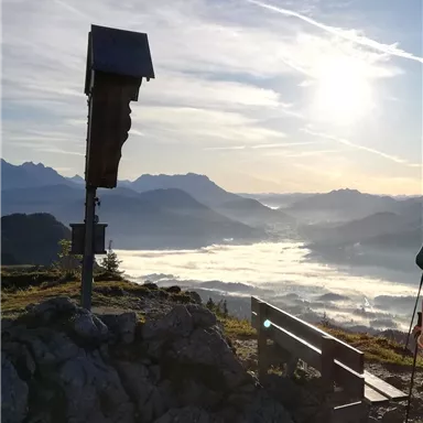 Ein Blick auf eine malerische Berglandschaft mit Nebel im Tal und der Sonne am Himmel. Im Vordergrund steht ein Holzbänkle und eine kleine Holzstruktur.