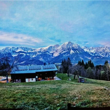 A picturesque alpine landscape with snow-covered mountains in the background. In the foreground, there is a cozy building on a green meadow.