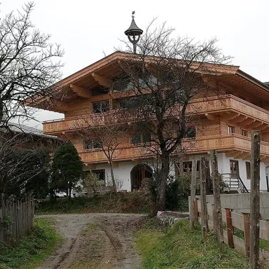 A traditional wooden house with several balconies and a characteristic roof. The garden is surrounded by trees, and a gravel path leads to the entrance.
