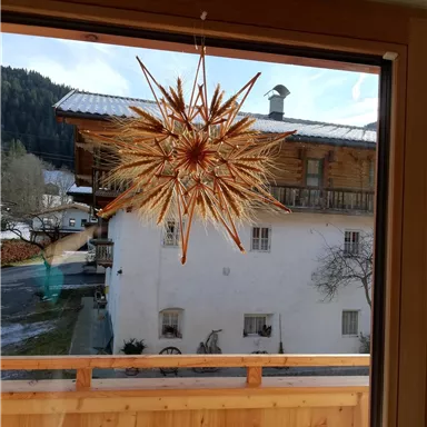 A beautifully decorated star hangs in front of a window. In the background, there is a traditional house and a picturesque winter landscape.