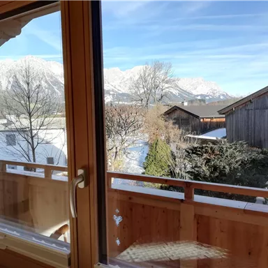 A view from a window onto snowy mountains and adjacent wooden houses. The sky is clear and blue.