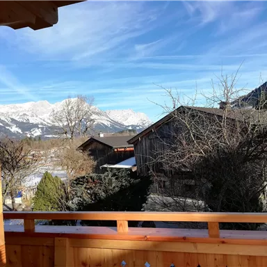 A view of the Alps with snow-covered mountains and traditional wooden houses. The sky is clear and blue with some clouds.