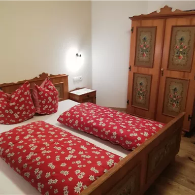 A cozy bedroom with a wooden bed and red pillows. In the background, there is a decorated wardrobe.