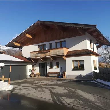 Ein modernes Haus im alpenländischen Stil mit einem großen Balkon. Es liegt in einer schneebedeckten Umgebung unter klarem Himmel.