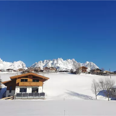 A picturesque winter landscape with snow-covered hills and majestic mountains in the background. The clear blue sky complements the tranquil atmosphere.