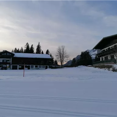 Eine winterliche Landschaft mit schneebedeckten Gebäuden. Im Hintergrund sind Bäume und Berge zu sehen.