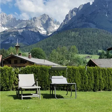 A relaxed garden landscape with two lounge chairs and a table. In the background, majestic mountains rise under a blue sky.