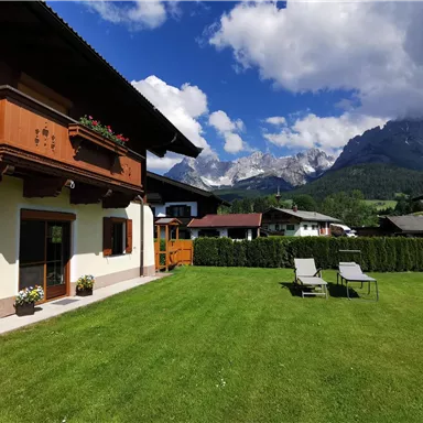 A cozy house with a colorful garden and a meadow. In the background, impressive mountains and a blue sky can be seen.
