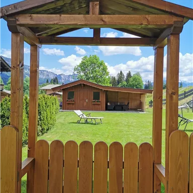 A wooden viewpoint overlooking a well-maintained garden and a hut. In the background, green meadows and a blue sky with clouds can be seen.