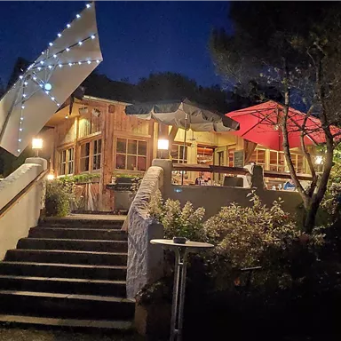Ein gemütliches Restaurant bei Nacht, beleuchtet von warmem Licht. Die Terrasse ist mit bunten Sonnenschirmen ausgestattet.