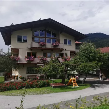 A charming building with blooming balcony boxes. In the foreground, flowerbeds and a playground can be seen.