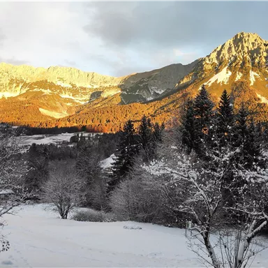 Eine verschneite Landschaft mit Bergen im Hintergrund. Die Sonne beleuchtet die Bergspitzen und verleiht der Szene ein warmes Licht.