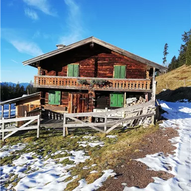 Eine traditionelle Hütte aus Holz mit grünen Fensterläden. Umgeben von Wiesen und Schnee unter einem klaren blauen Himmel.
