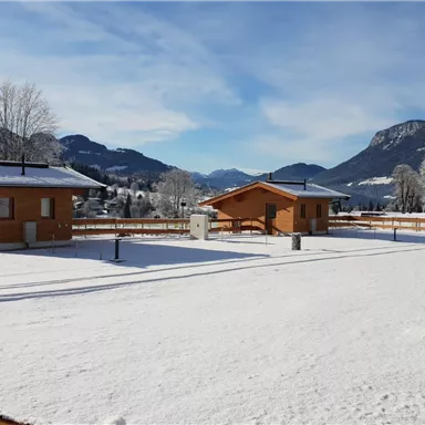 Winter landscape with wooden huts and snow-covered ground. In the background, mountains and a clear sky are visible.