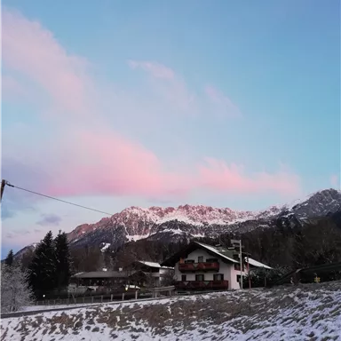 Eine winterliche Landschaft mit schneebedeckten Bergen und einem hübschen Haus. Der Himmel zeigt sanfte rosa und blaue Farbtöne.