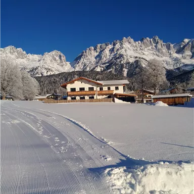 A picturesque winter landscape with snow-covered fields and a cozy house. In the background, majestic mountains rise under a clear blue sky.
