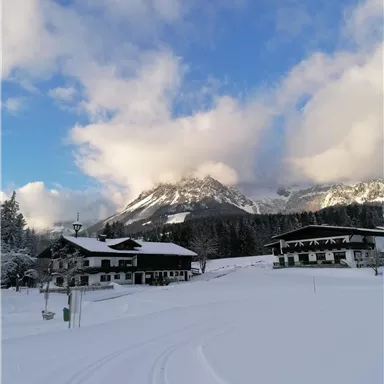 Eine winterliche Landschaft mit schneebedeckten Hügeln und Bergen im Hintergrund. Im Vordergrund stehen traditionelle Holzgebäude unter einem klaren Himmel.