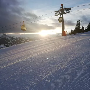Eine Skilandschaft mit frisch präparierter Piste und einem Seilbahn-Untergestell. Der Sonnenuntergang lässt die Szene in warmen Farben erstrahlen.