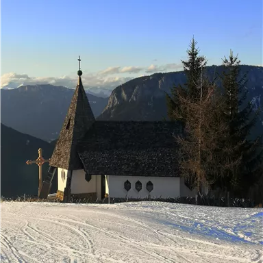 A small church in a snowy landscape with mountains in the background. The sky is clear and blue.