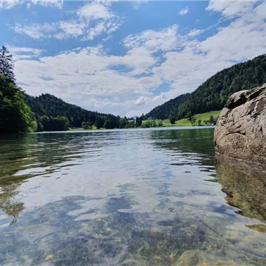 A tranquil lake with clear water and gentle waves. Surrounded by green hills and wooded shores under a blue sky.