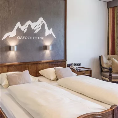 Ein gemütliches Schlafzimmer mit zwei Betten und schönen Holzmöbeln. An der Wand ist eine Bergsilhouette mit dem Schriftzug „Oafach Hetzig“ zu sehen.