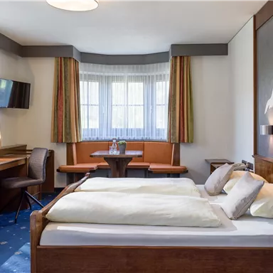 Ein gemütliches Zimmer mit einem großen Bett, einem Tisch und Stühlen. Helle Fenster sorgen für natürlichem Licht und eine angenehme Atmosphäre.