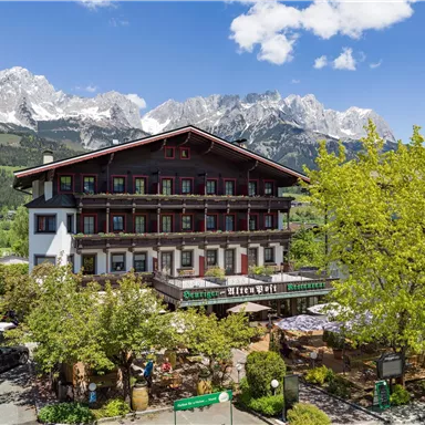 Ein charmantes Hotel mit traditioneller Architektur, umgeben von grünen Bäumen. Im Hintergrund erheben sich majestätische Berge unter einem klaren Himmel.